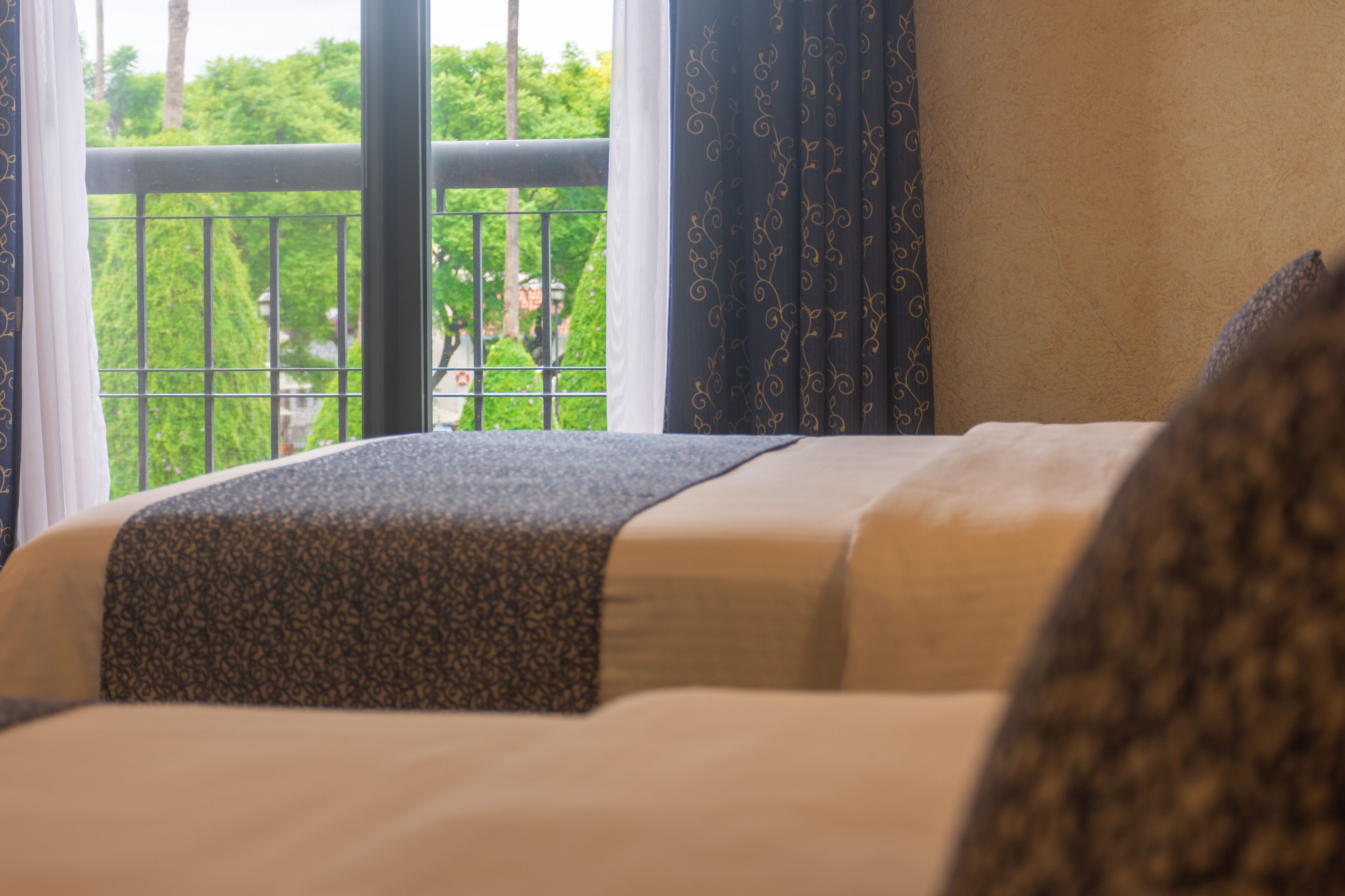 A hotel room with a bed, a view of greenery outside, and a window with curtains.