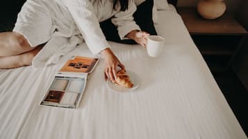 Une personne assise sur un lit, avec un livre, une assiette contenant un croissant et une tasse.