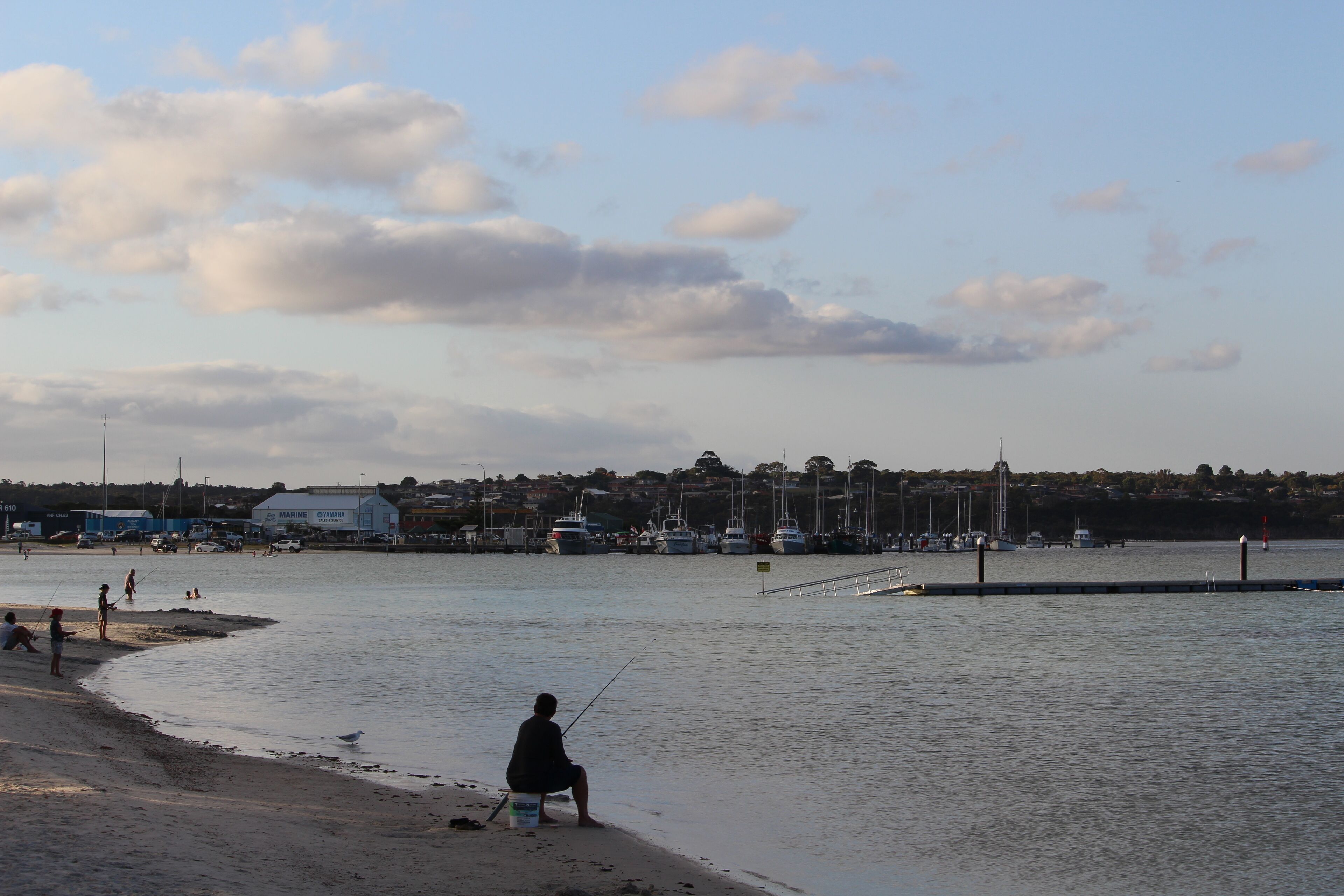 Une plage où une personne pêche, avec des bateaux en arrière-plan et un port de plaisance avec des bâtiments.