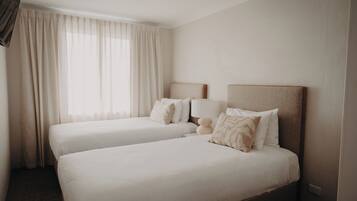A hotel room with two beds, a large window with curtains, a flat-screen TV, and a bedside table with a lamp.