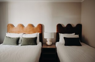 Una habitación de hotel con dos camas, cada una con un diseño único de cabecera, una mesita de noche con una lámpara y una pequeña estantería debajo de la mesa.