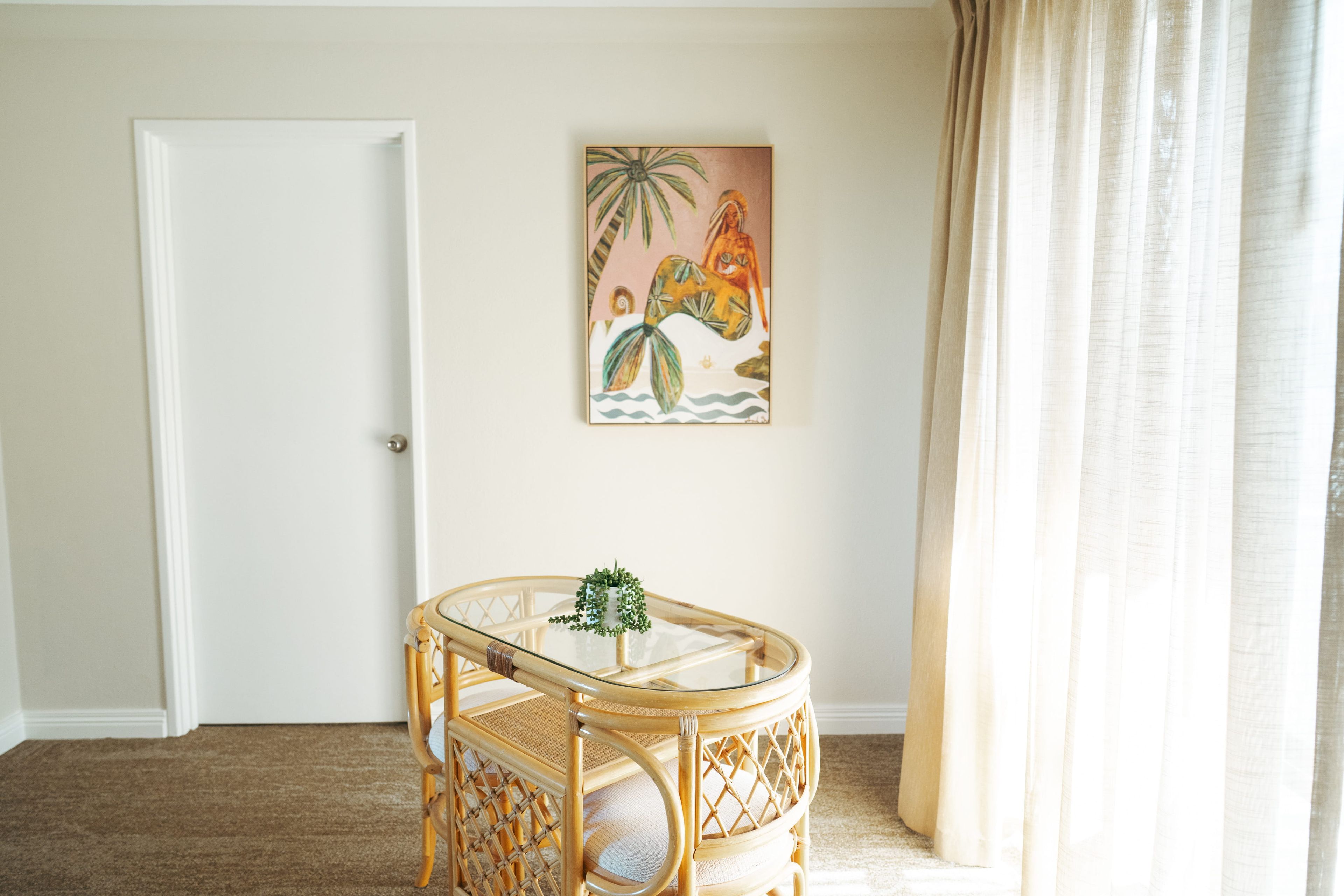 A room with a door, a glass-top table with a plant, a wicker chair, and a painting on the wall.