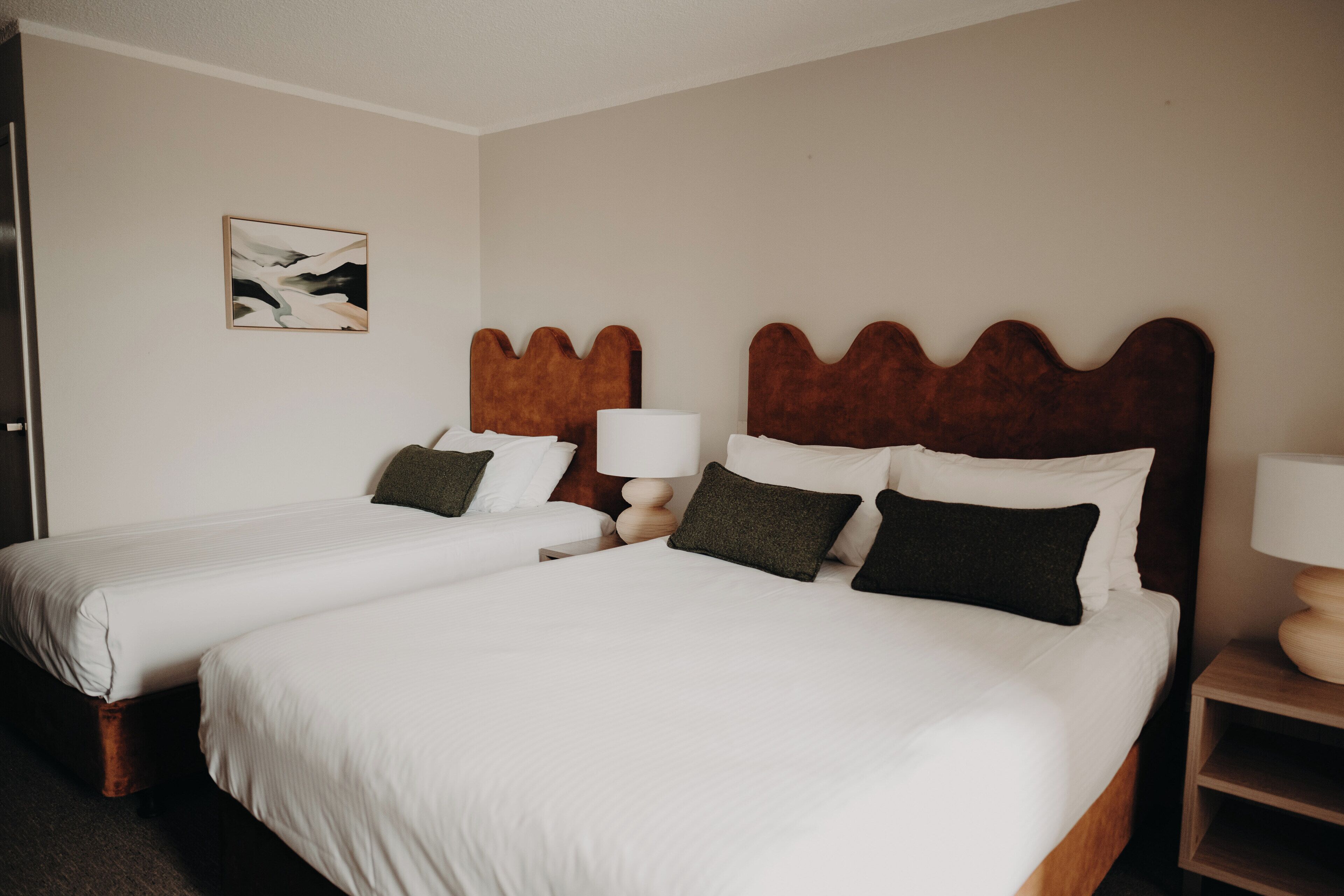 Habitación de hotel con dos camas, cabecera de madera, mesita de noche con lámpara y una obra de arte colgada en la pared.