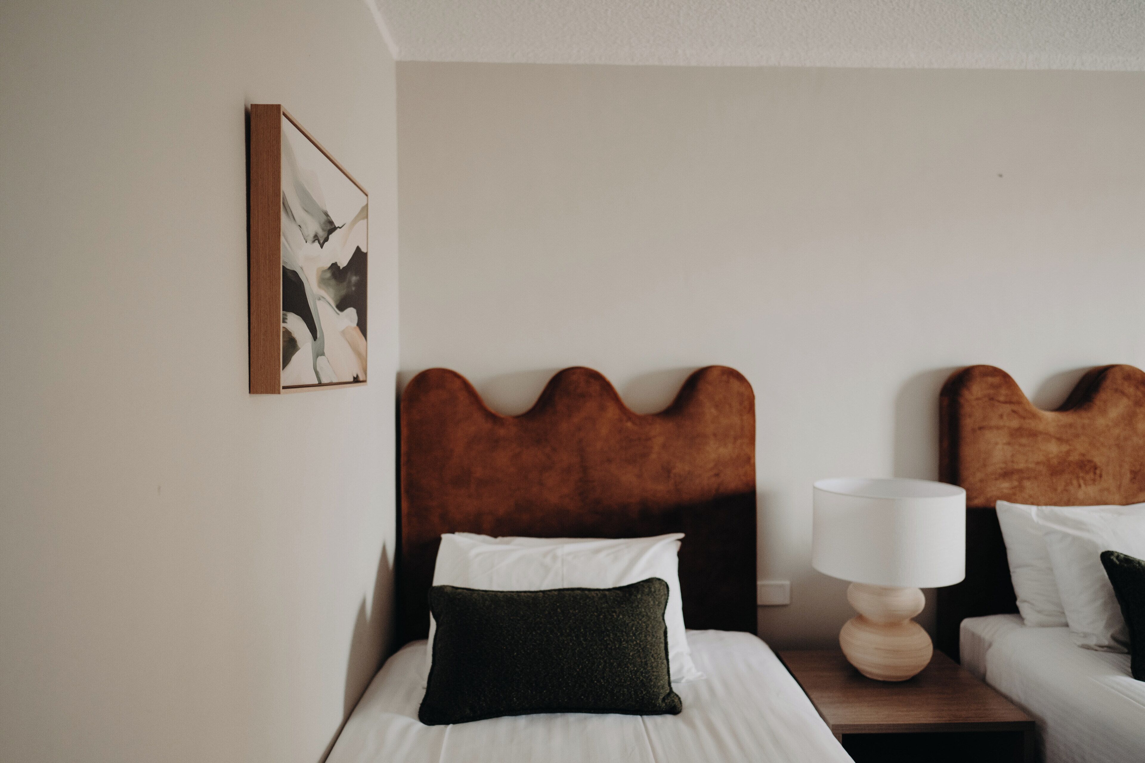 Ein ordentlich bezogenes Bett mit Holz-Kopfteil, ein Nachttisch mit Lampe und ein gerahmtes abstraktes Gemälde an der Wand.