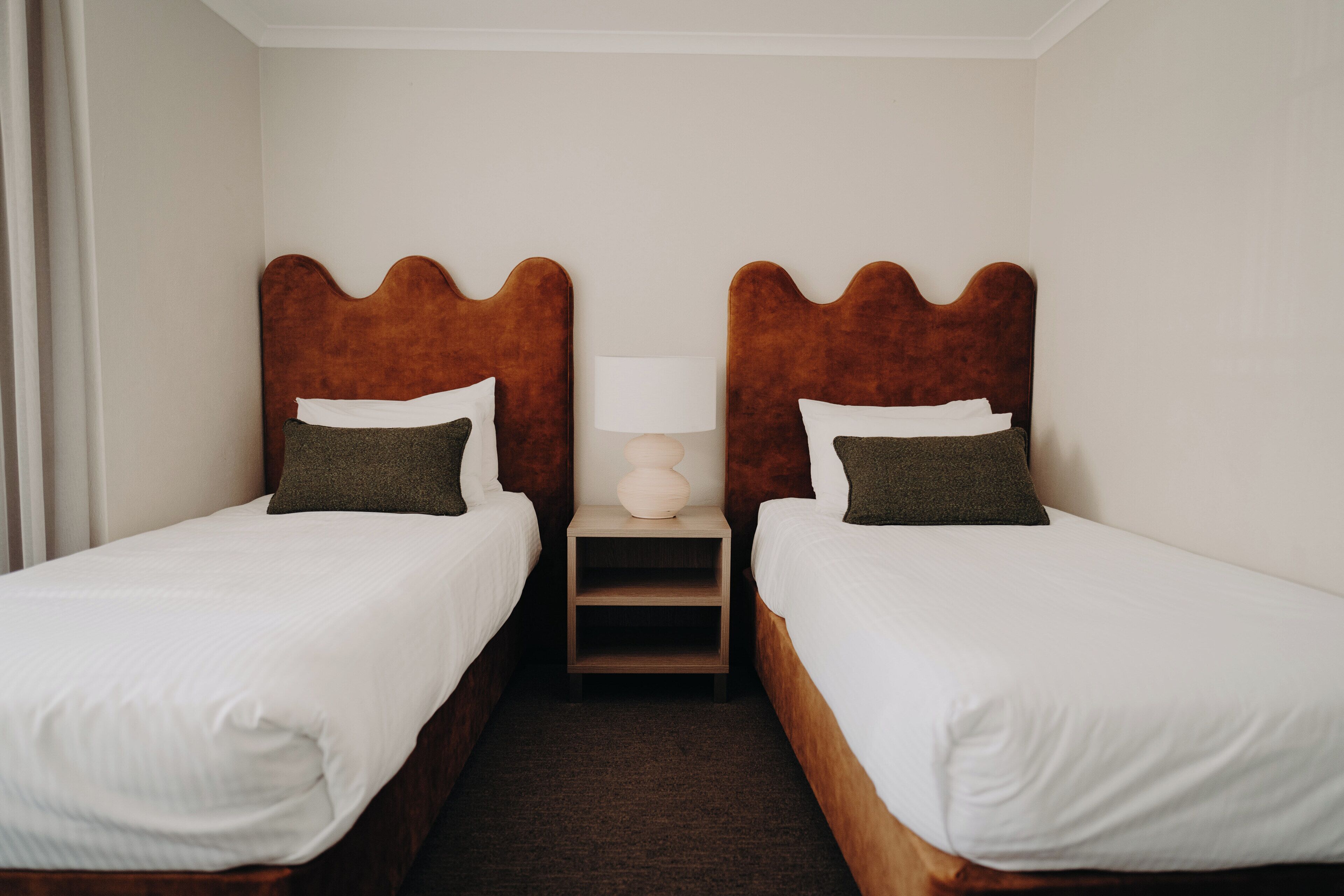 A hotel room with two single beds, a nightstand with a lamp, and a small wooden shelf.