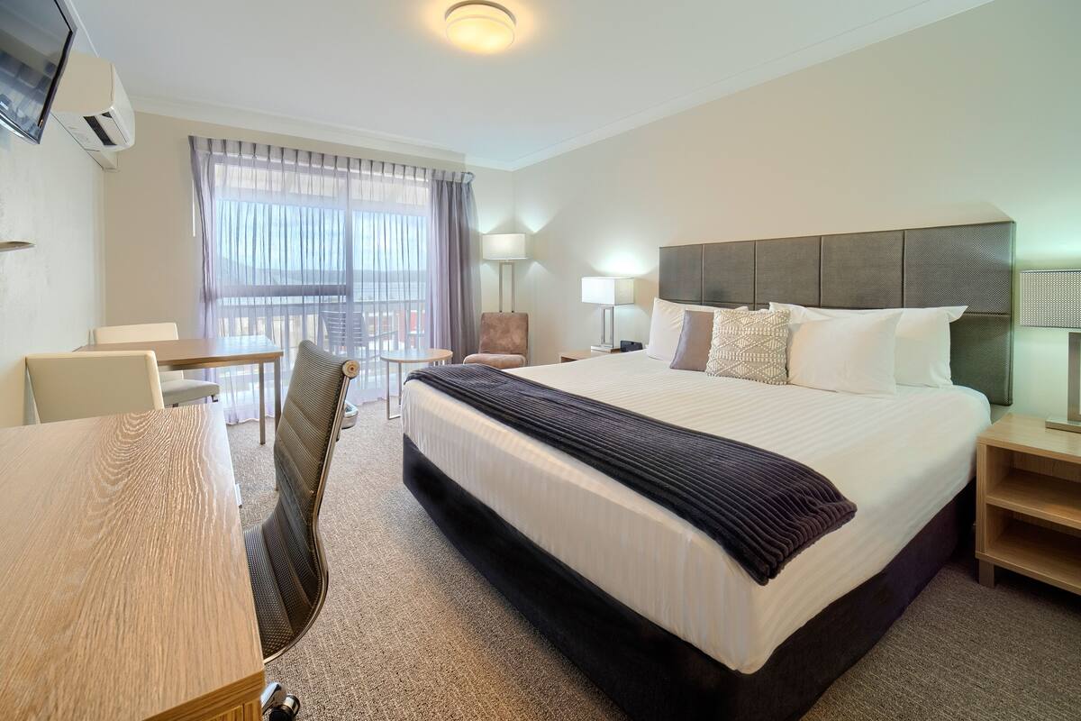 Ein Hotelzimmer mit einem großen Bett, einem Schreibtisch, einem Stuhl, einem Fernseher und einem Vorhangfenster.