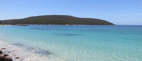 A beach with clear turquoise water, rocky shoreline, and a distant green hill.