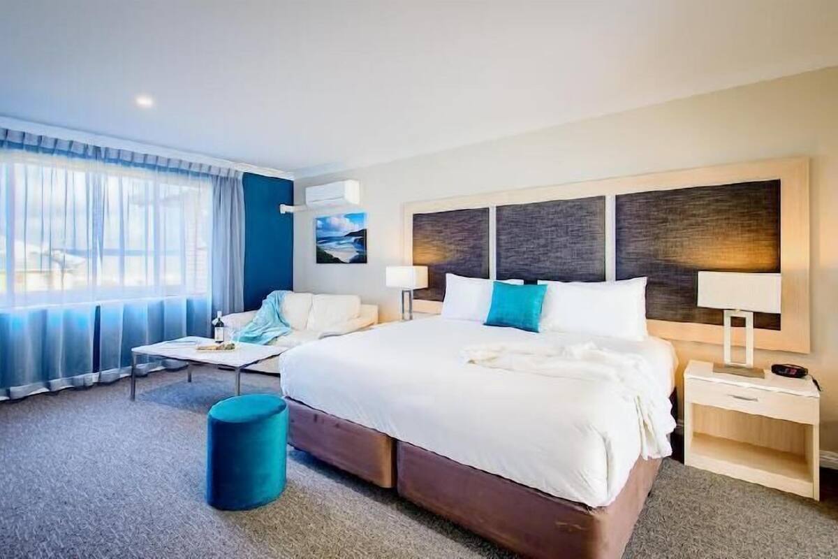 Ein Hotelzimmer mit einem großen Bett, einem Nachttisch, einem Sessel, einem kleinen Tisch und einem Fenster mit Vorhängen.