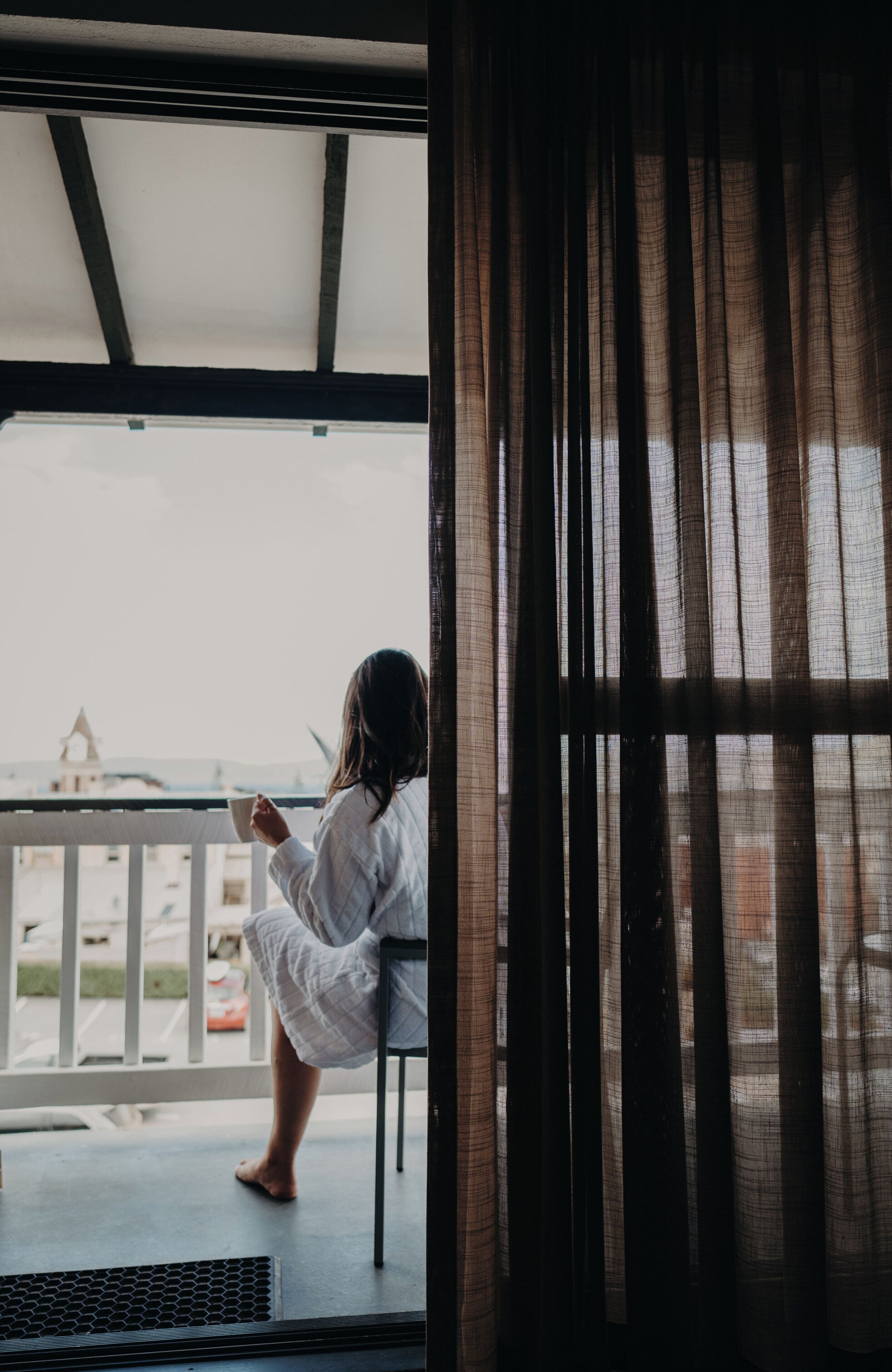 A person sitting on a chair by a window with a view of a building and a balcony.