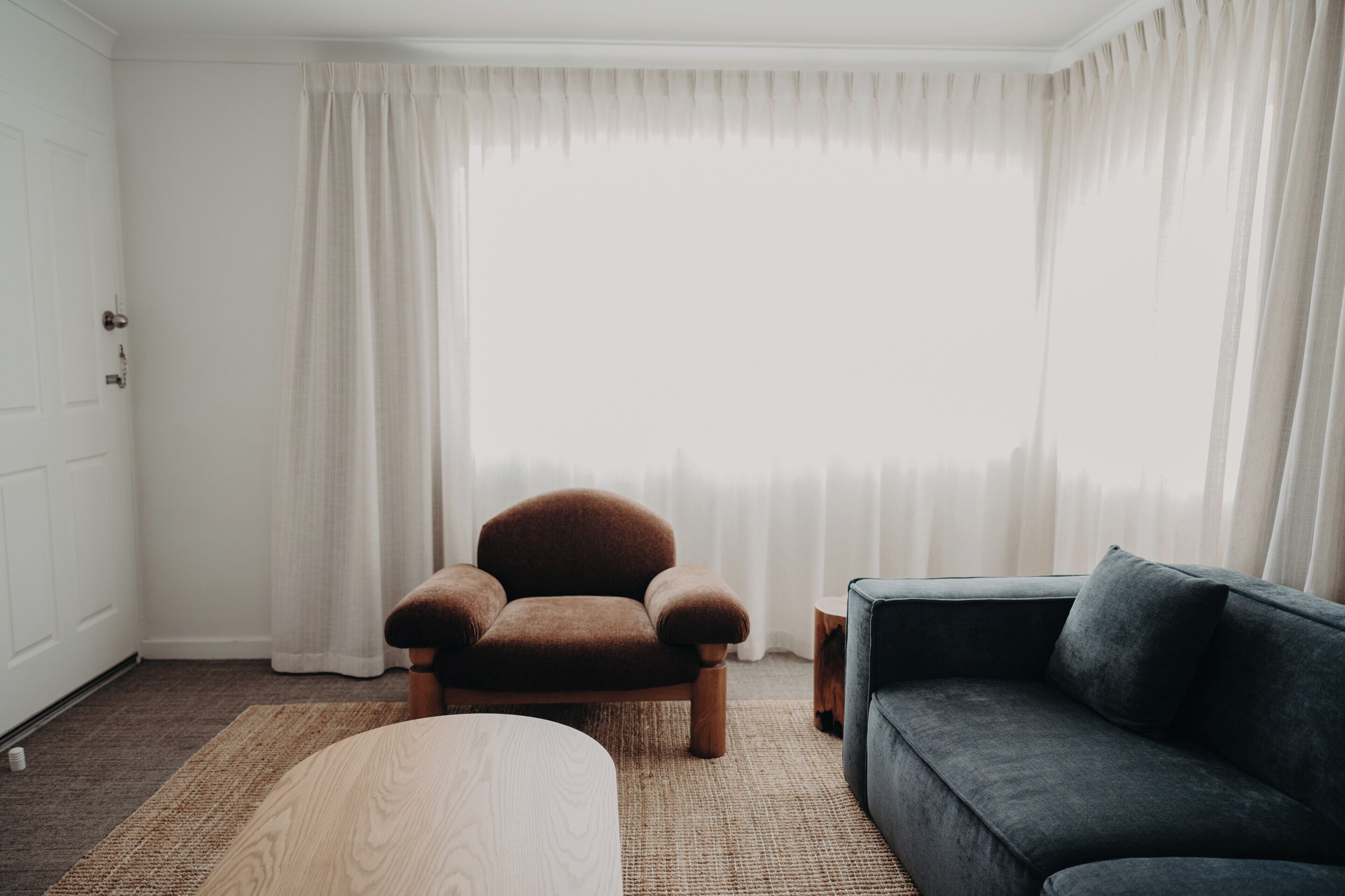 Una habitación con un sillón marrón, un sofá oscuro, un reposapiés de madera y un ventanal con cortinas blancas.
