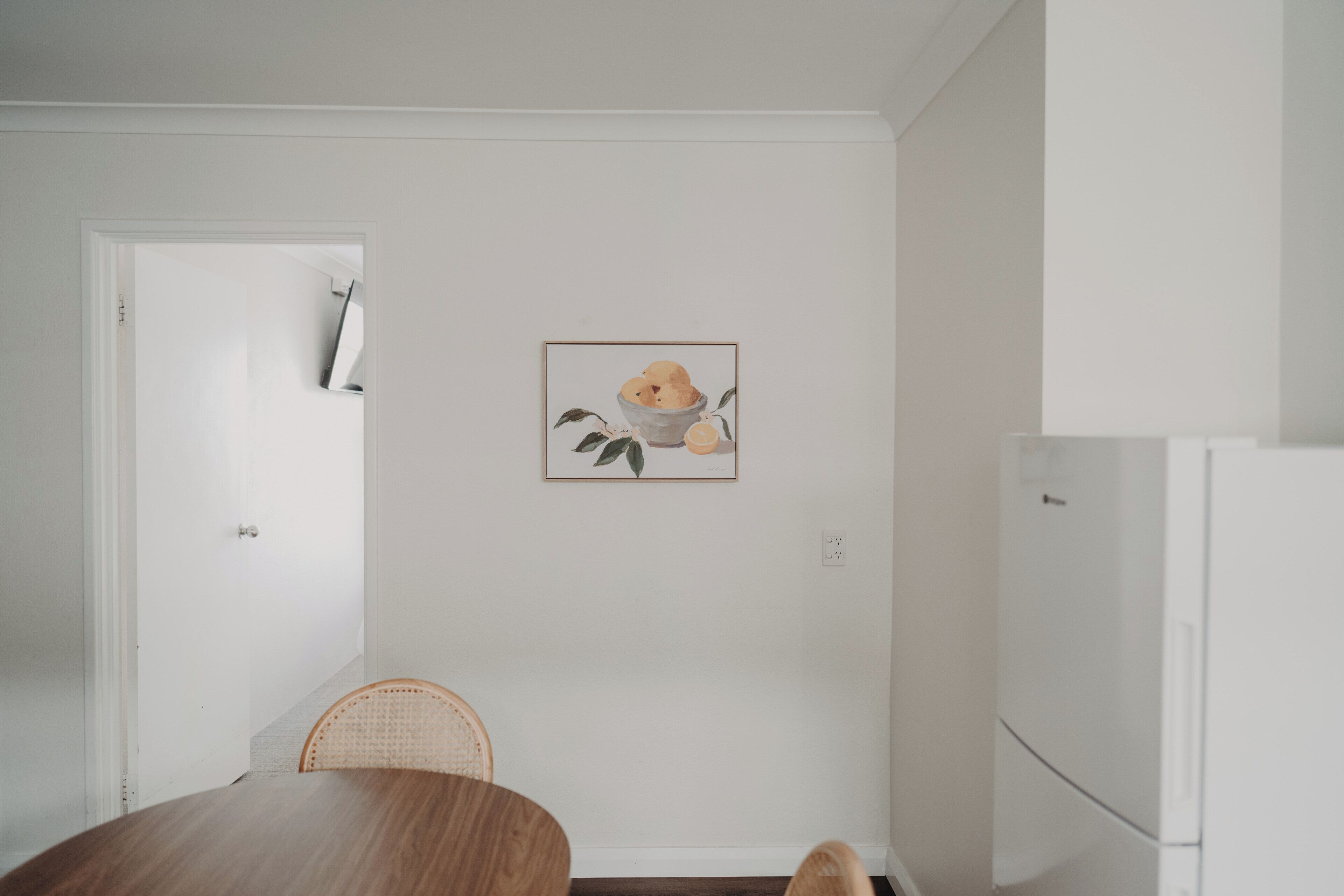 A dining area with a wooden table, a wicker chair, a white wall with a framed picture, and a white refrigerator.