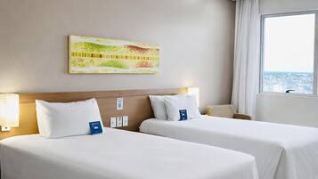 A hotel room with two beds, a large window, a ceiling-mounted security camera, and a wall-mounted artwork.