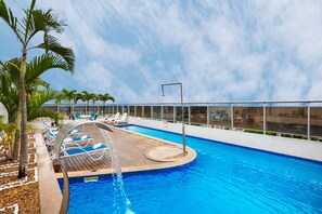 Uma piscina com deck de madeira, cadeiras de descanso e palmeiras.