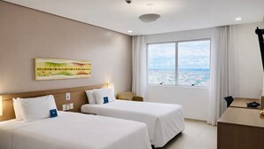 Ein Hotelzimmer mit zwei Betten, einem Schreibtisch, einem Stuhl, einem großen Fenster mit Blick auf die Stadt und einer Deckenkamera.