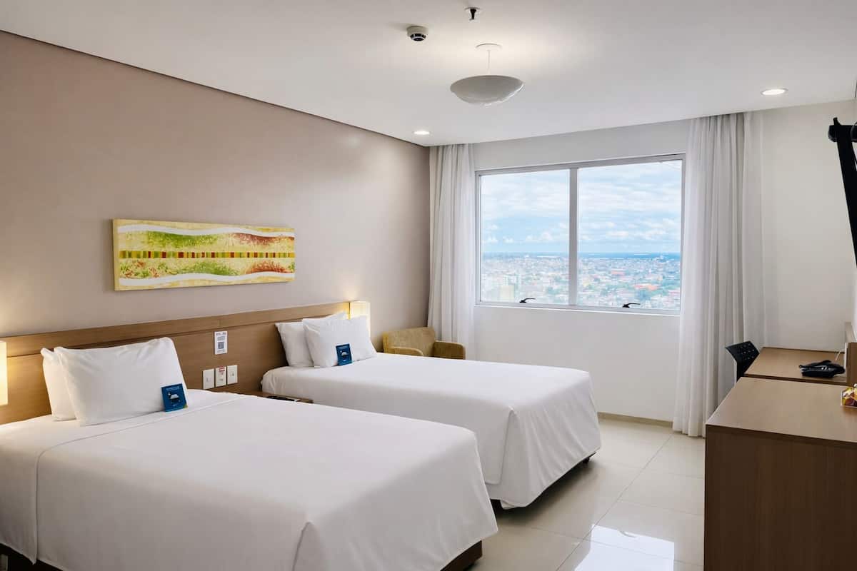 Habitación de hotel con dos camas, un escritorio, una silla, un ventanal con vistas a la ciudad y una cámara de seguridad instalada en el techo.