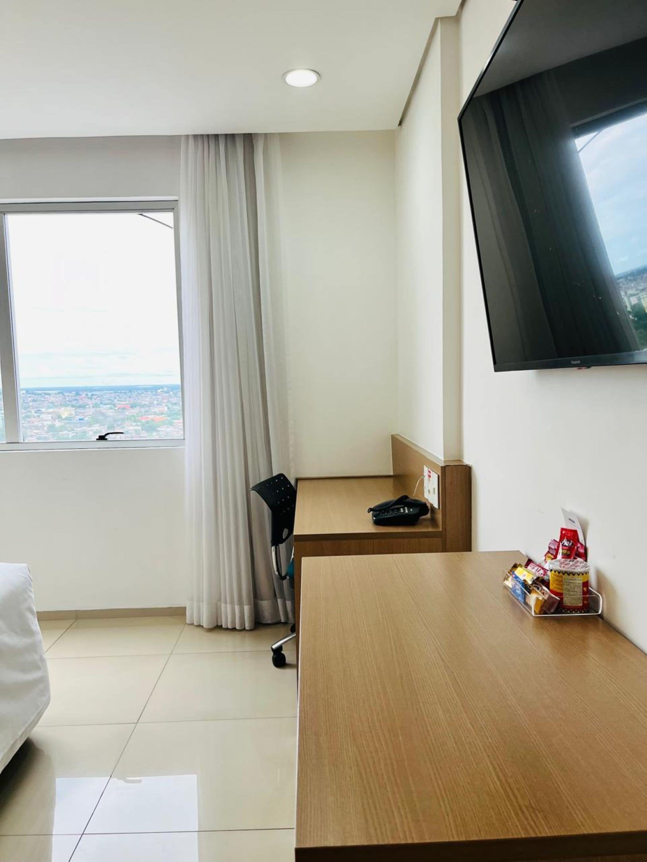 Habitación de hotel con cama, escritorio con silla, televisor, ventana con cortinas y vista panorámica de la ciudad.