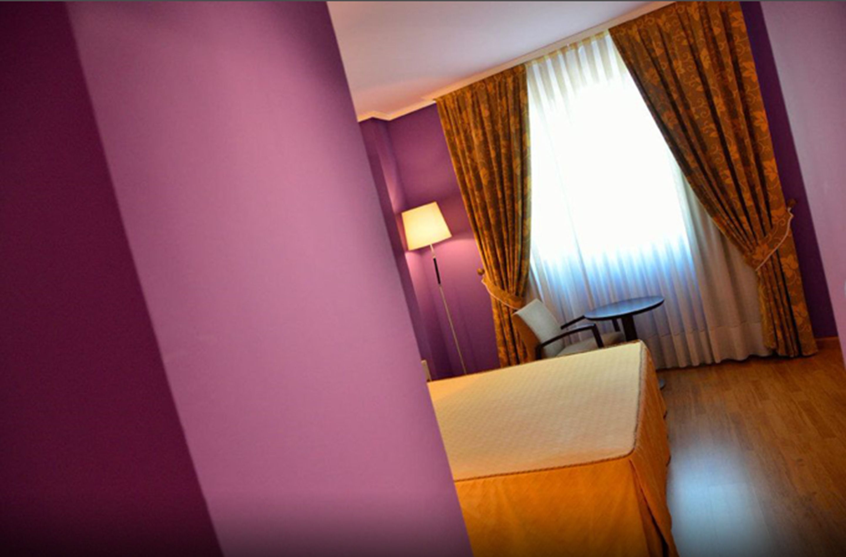 Habitación de hotel con paredes moradas, cama con cubierta color beige, una mesita, una lámpara y una ventana con cortinas.