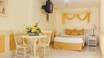 Una habitación de hotel con cama, mesita de noche, lámpara, televisor montado en la pared y una mesa con sillas y un jarrón de flores.