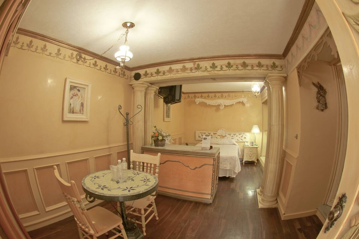 Ein Zimmer mit rundem Tisch, Stühlen, einem Bett, einem Nachttisch und einem kleinen Schreibtisch mit Lampe.
