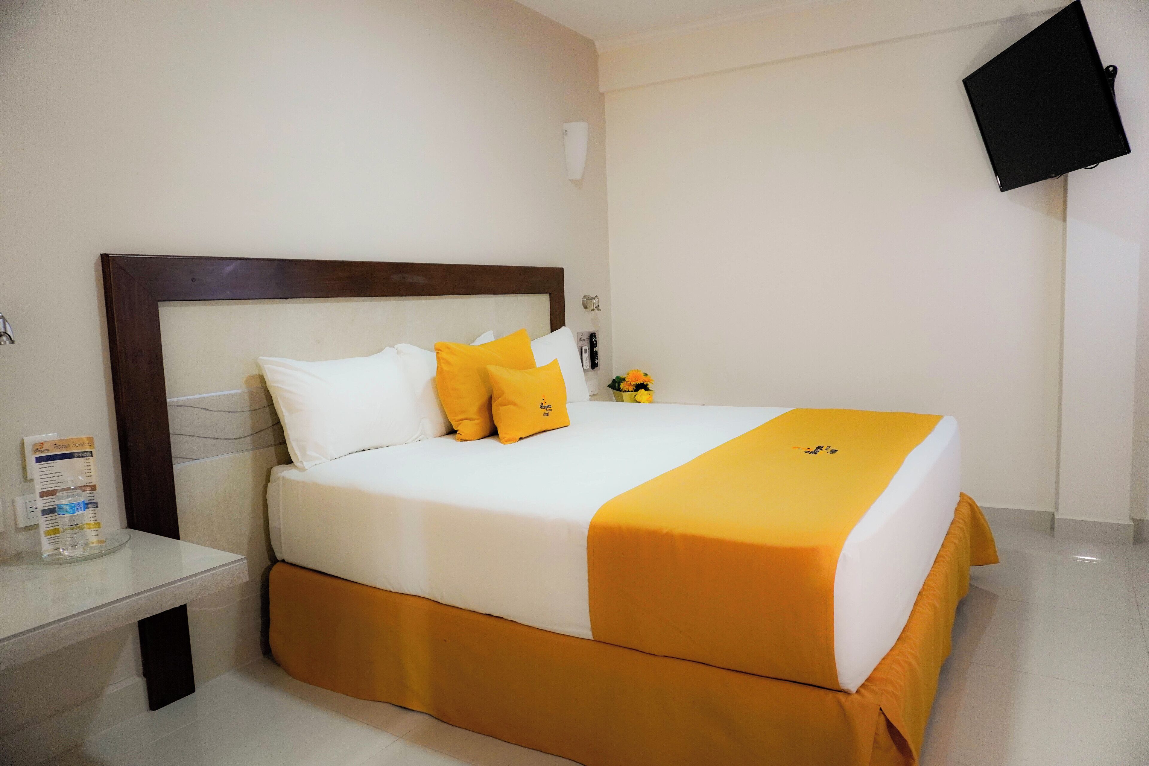 A hotel room with a large bed, a bedside table, a wall-mounted TV, and a small table with a vase of flowers.