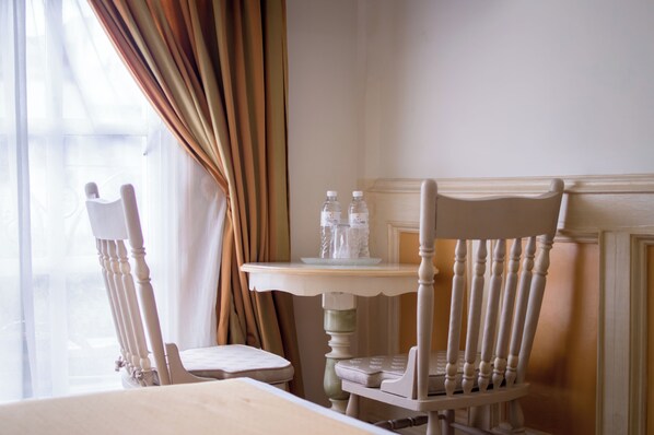 Una habitación con dos sillas blancas, una mesita con botellas de agua y una ventana con cortinas.