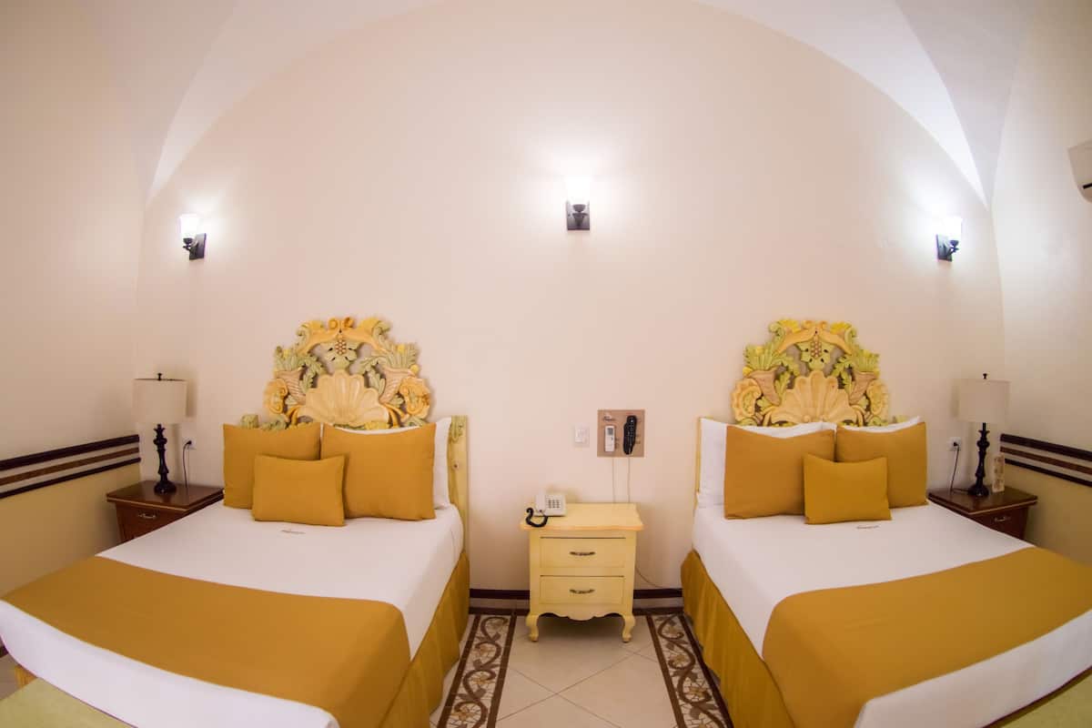 Une chambre d’hôtel avec deux lits, chacun doté d’une tête de lit ornée de motifs floraux, et une petite table de chevet avec une lampe.