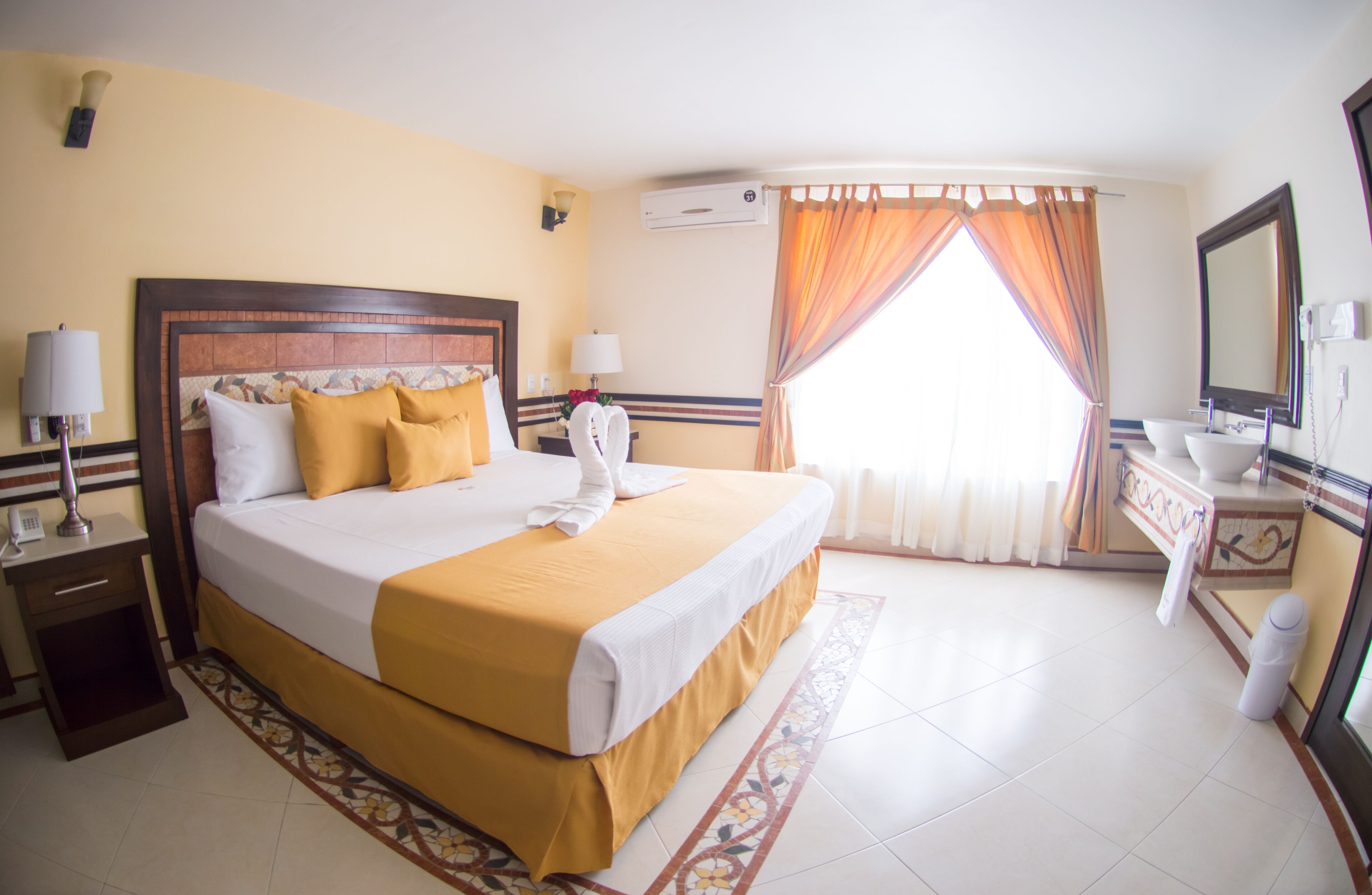 A hotel room with a large bed, a bedside table with a lamp, a bathroom with a sink and mirror, and a window with curtains.