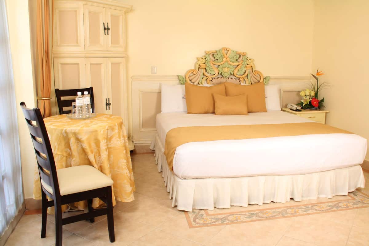Une chambre d’hôtel avec un grand lit, une chaise, une table avec des bouteilles d’eau et une table de chevet avec un téléphone et des fleurs.
