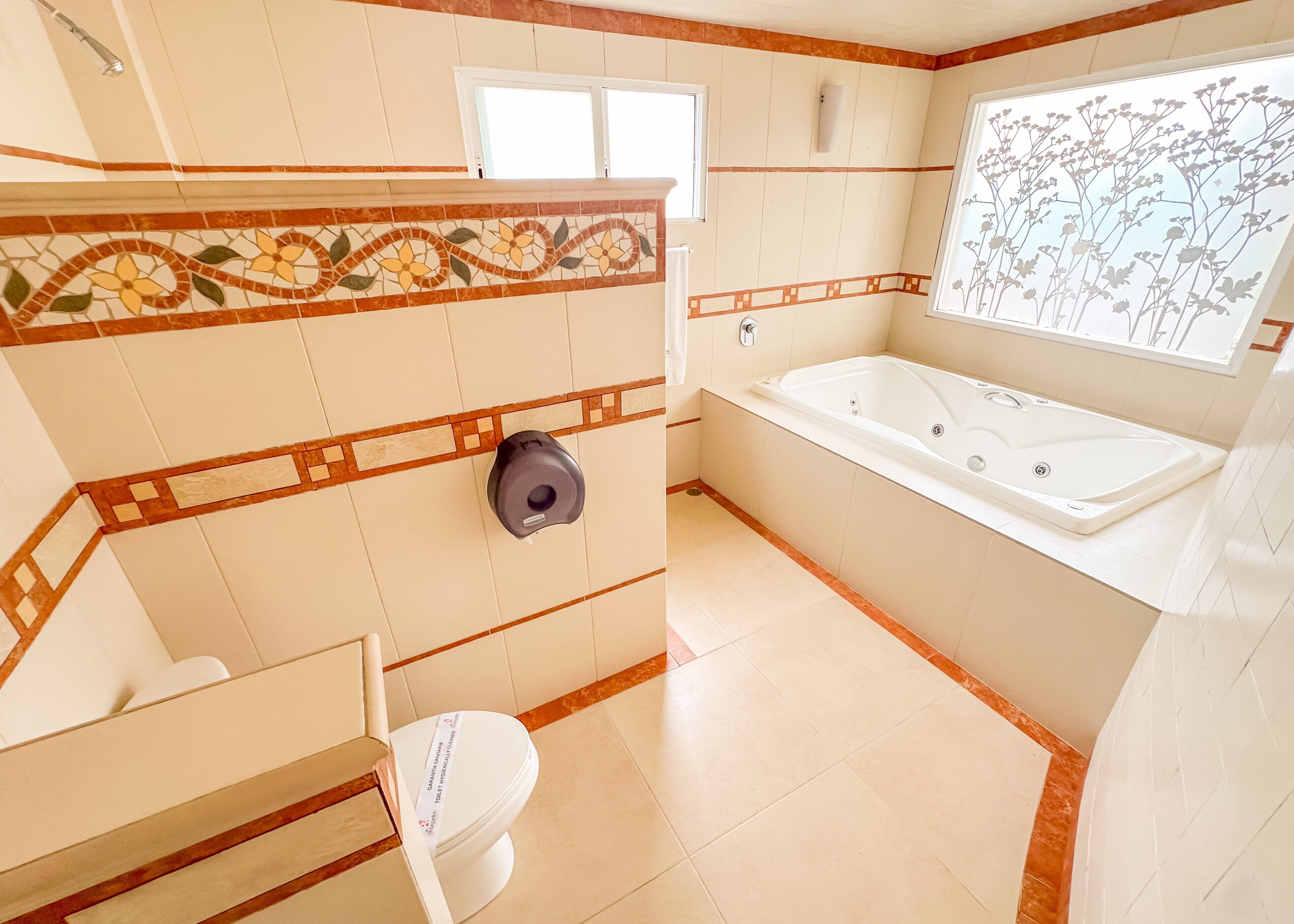 Un baño con inodoro, secamanos, bañera y ventana con vistas a un jardín floral.