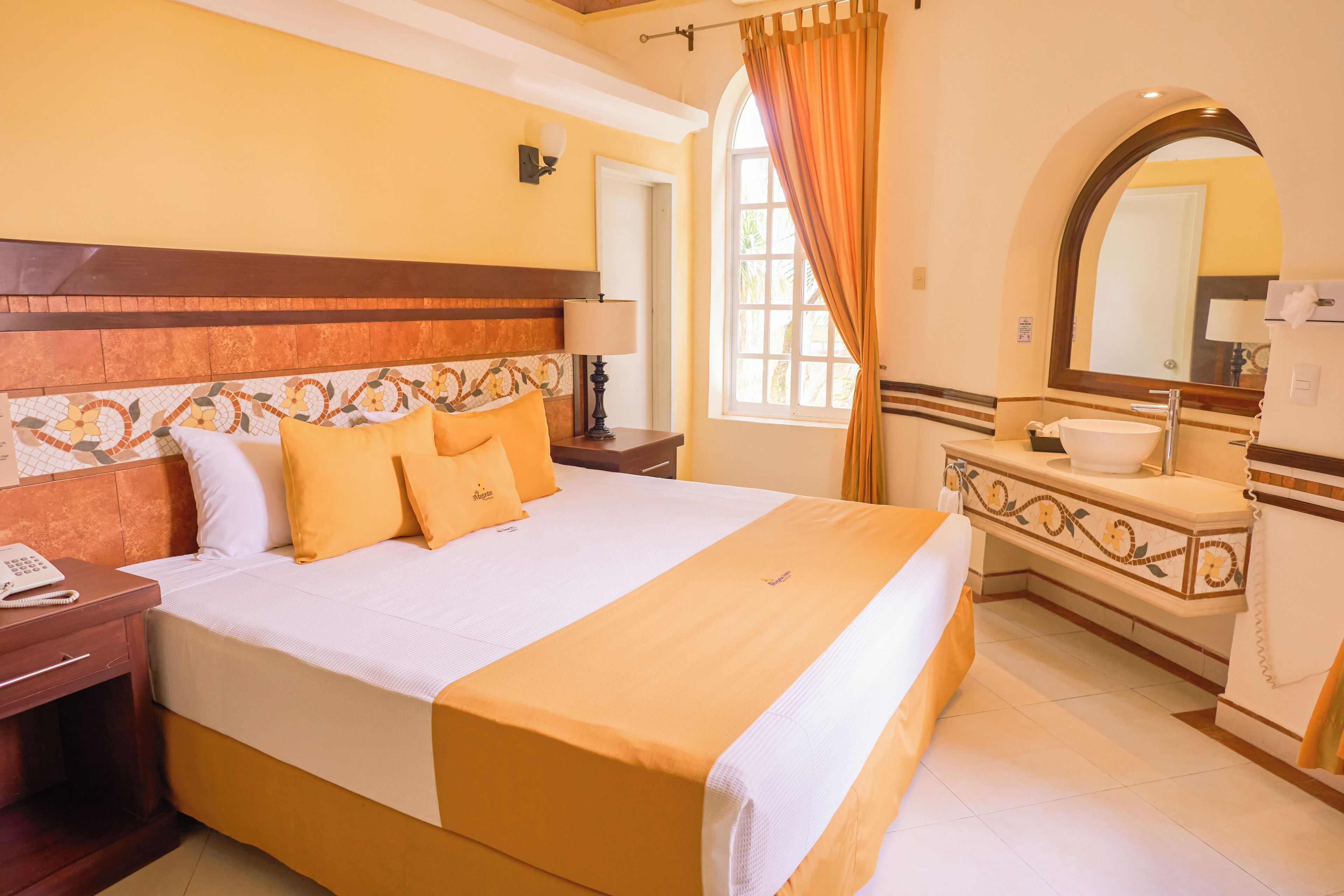 A hotel room with a large bed, a bedside table with a lamp, a bathroom with a sink and mirror, and a window with curtains.