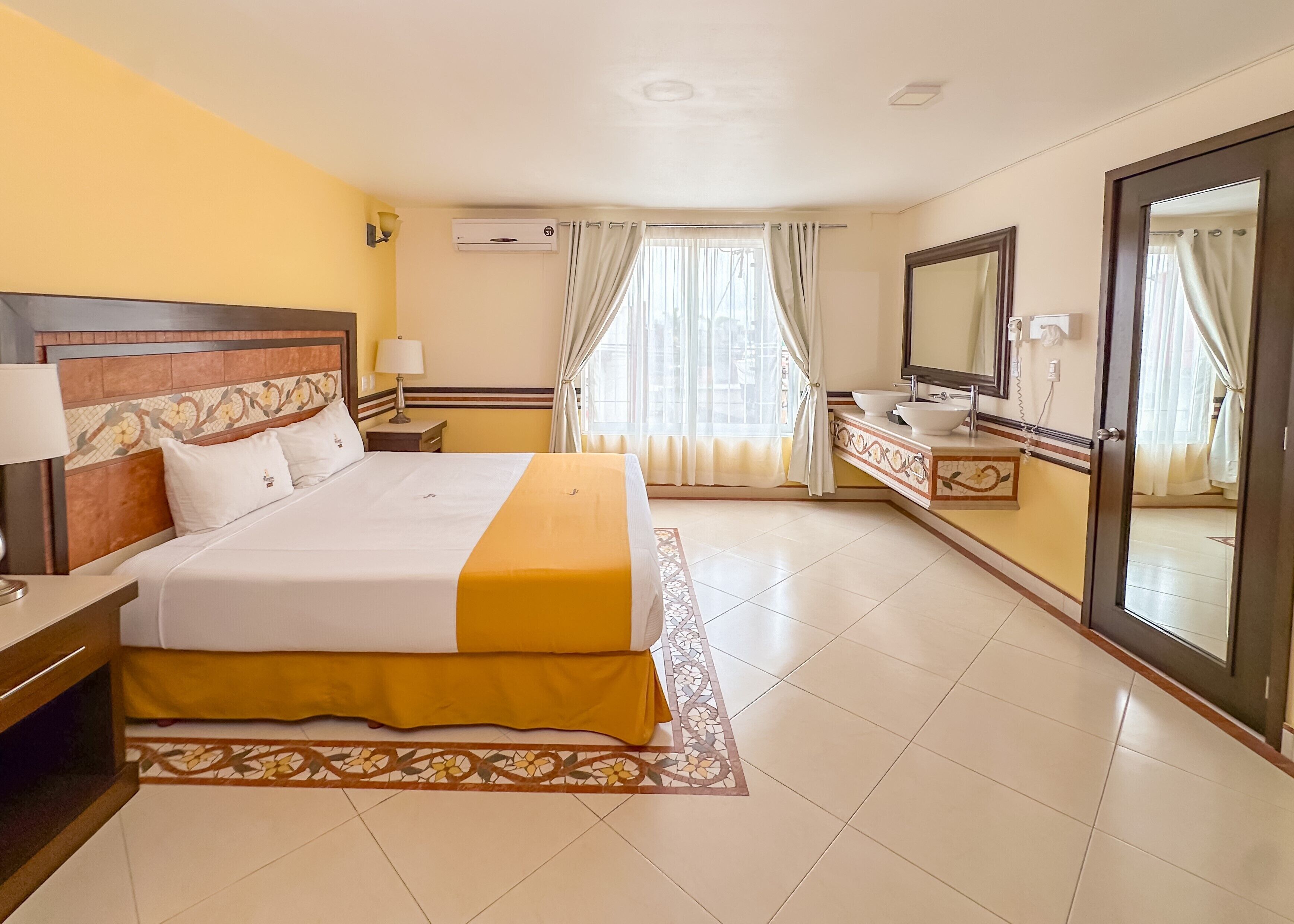 Une chambre d’hôtel comprenant un lit, une table de chevet, une lampe, un appareil de climatisation, un miroir dans la salle de bain, ainsi qu’une salle de bain avec un lavabo et des toilettes.