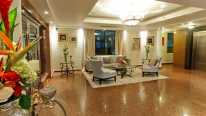 Een ruime lobby met een gepolijste vloer, een zithoek met een bank en fauteuils, een glazen salontafel en een kroonluchter.