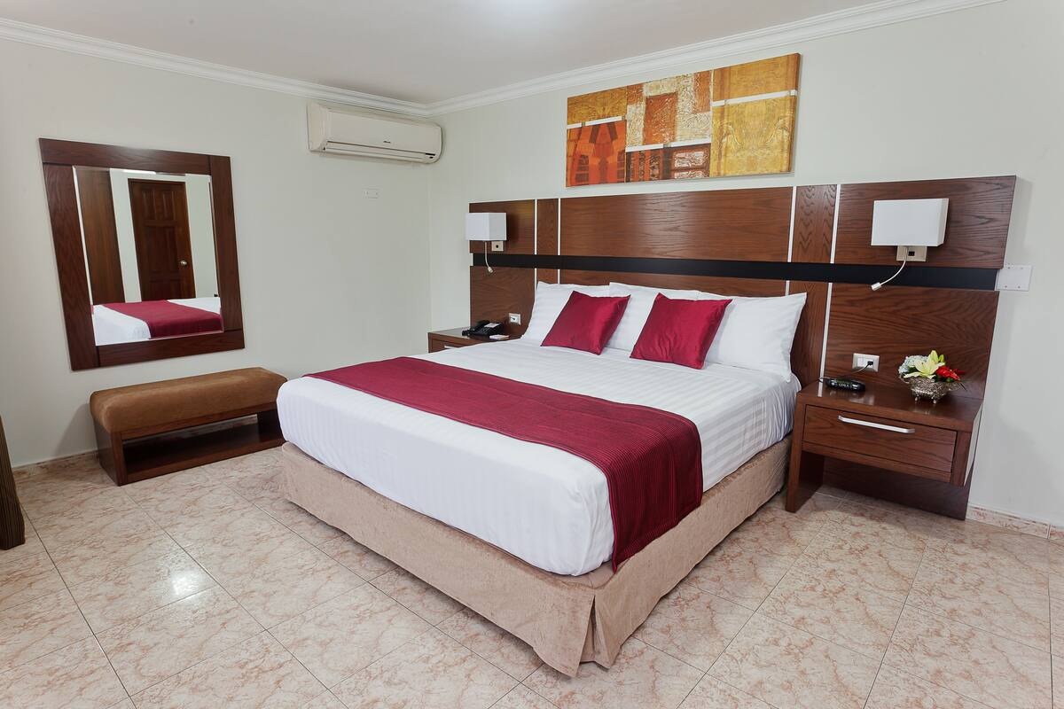 A hotel room with a large bed, a wooden headboard, a nightstand, a bench, a mirror, and a wall-mounted air conditioner.