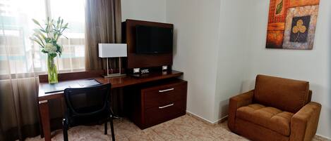 A hotel room with a desk, chair, TV, sofa, and artwork on the wall.