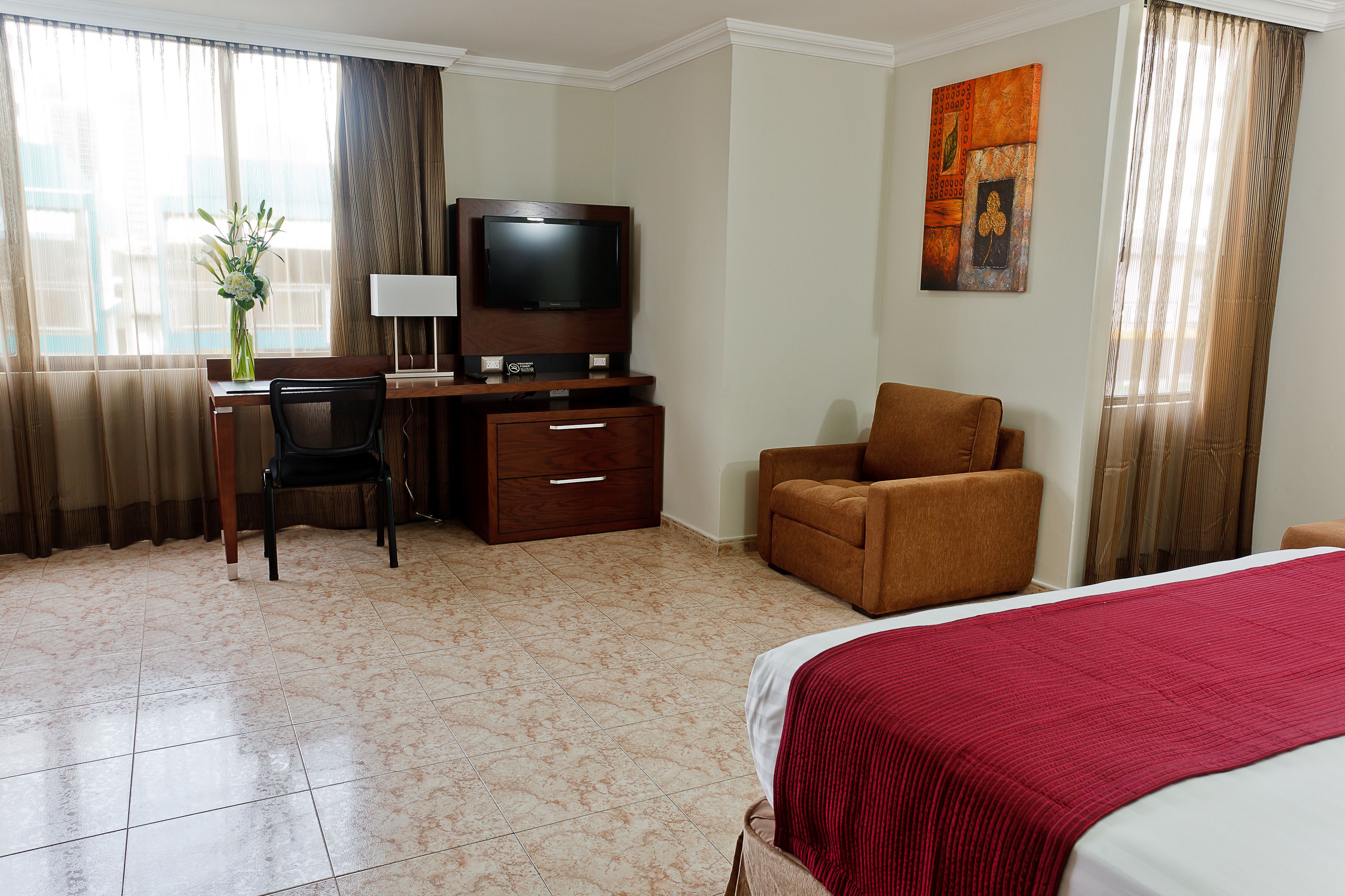 A hotel room with a bed, a desk with a chair, a television, a brown armchair, and a painting on the wall.