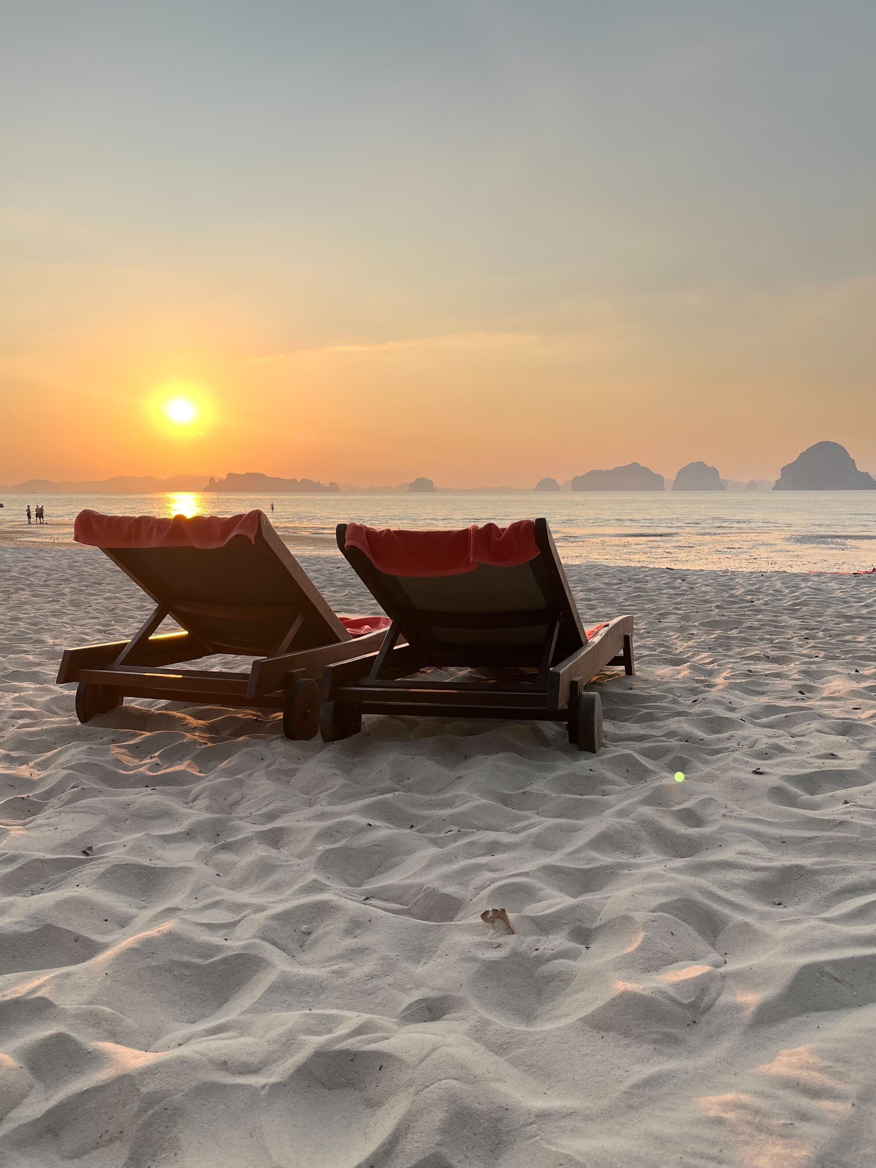 Dos sillones de madera con cojines rojos sobre una playa arenosa al atardecer.