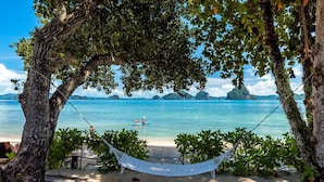 Una hamaca bajo un árbol en una playa arenosa, con vista al mar y a islas distantes.