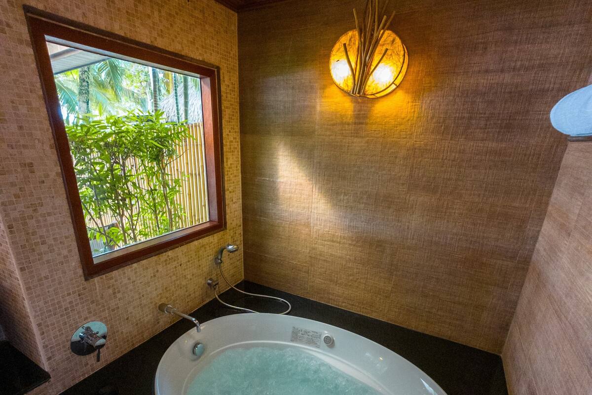 A bathroom with a bathtub, a window with a view of greenery, and a wall-mounted light fixture.