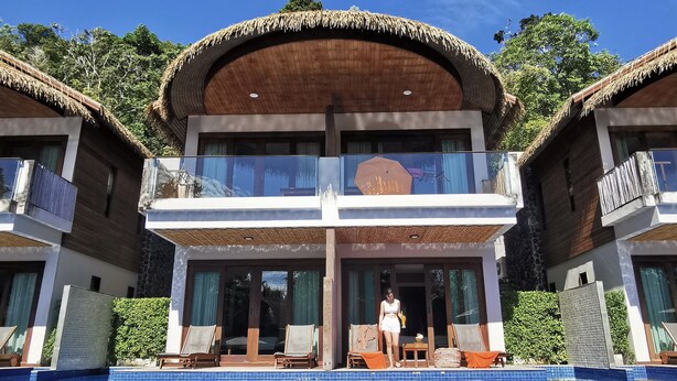 Un resort de dos pisos con piscina, un espacio al aire libre cubierto y una estructura con techo de paja.