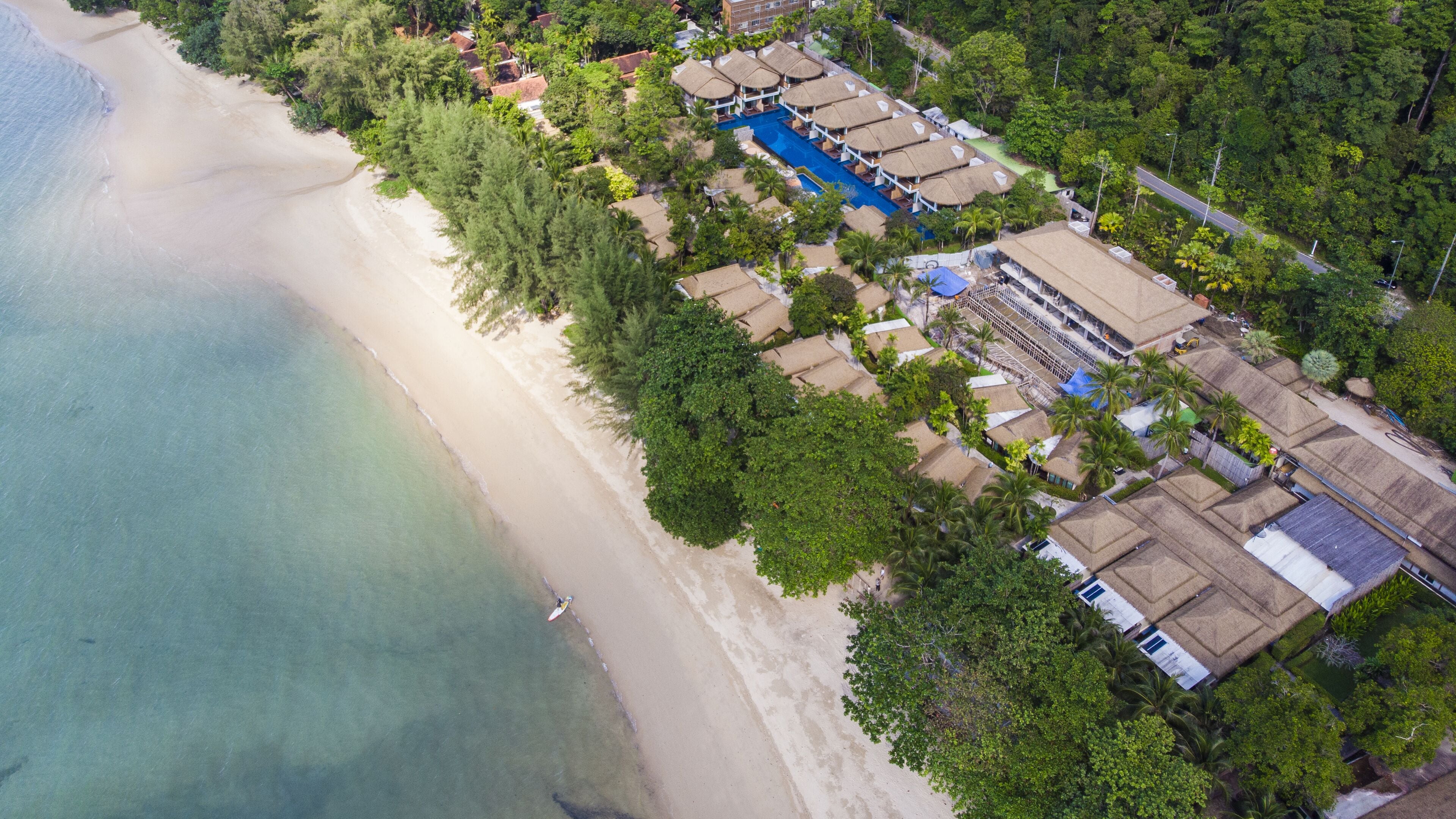 Vue aérienne d’un complexe hôtelier situé en bord de mer, avec une piscine, des bâtiments à toits de chaume et une végétation luxuriante.