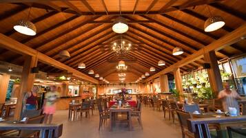 A spacious restaurant with wooden beams and chandeliers. Tables are arranged in the center, surrounded by chairs.