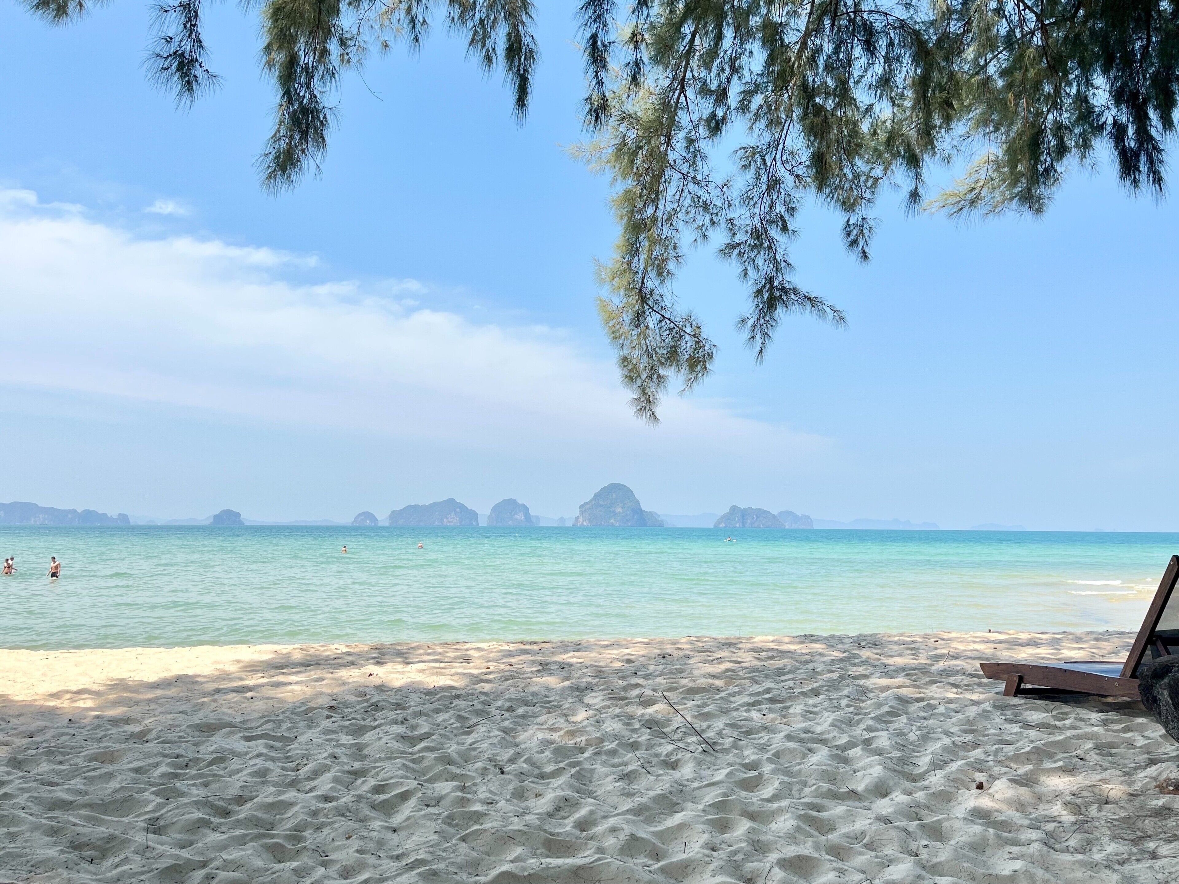 Une plage à l’eau turquoise et limpide, un rivage sablonneux, et quelques personnes qui se baignent.