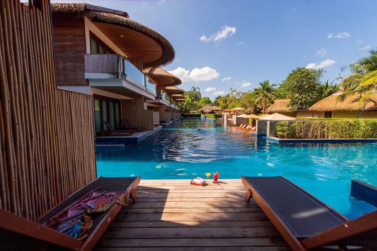 A pool area with wooden deck loungers, thatched-roof structures, and a clear blue sky.