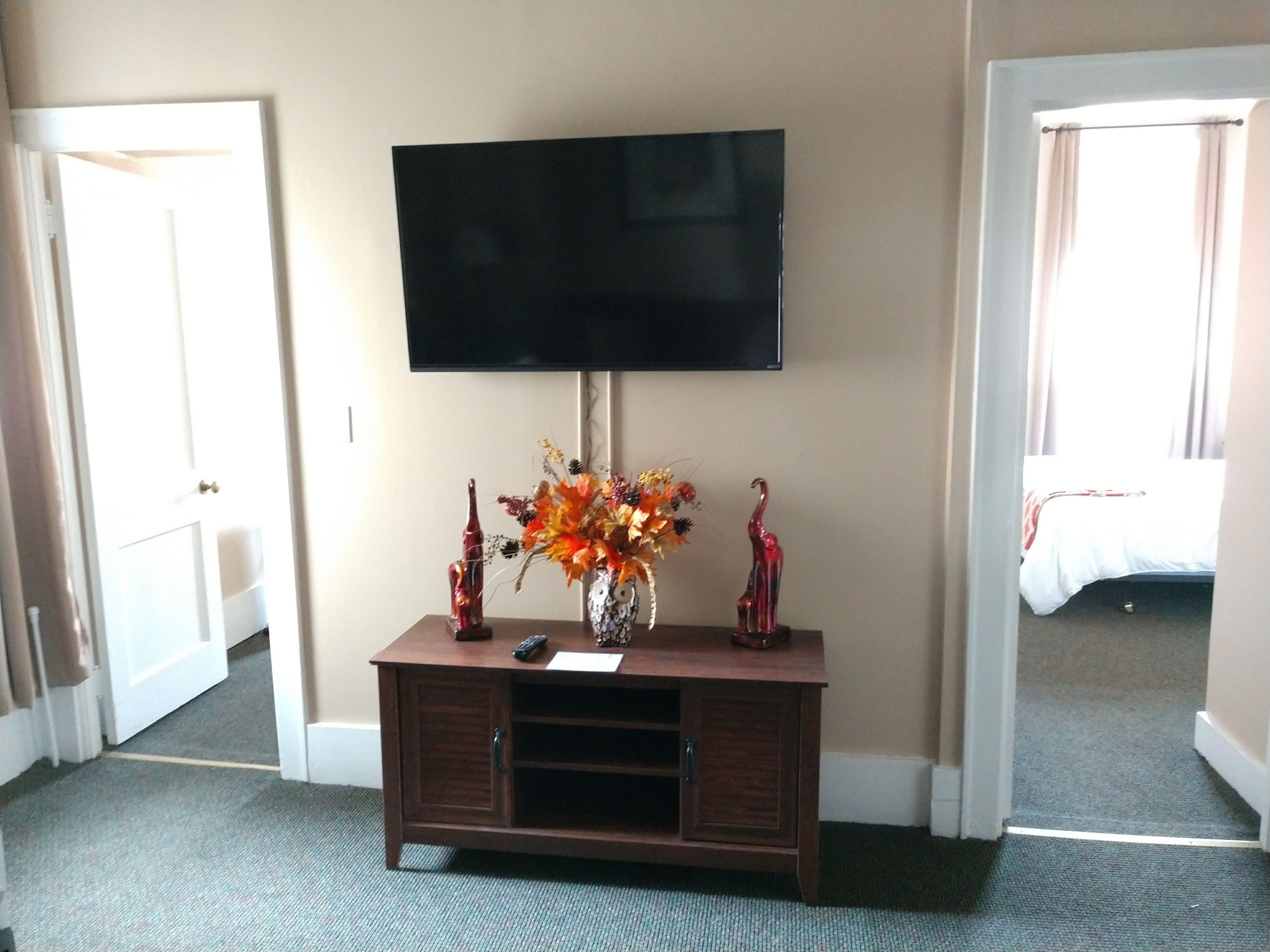 A hotel room with a flat-screen TV mounted on a wooden console, a vase with autumn flowers, and a bed with a white comforter.