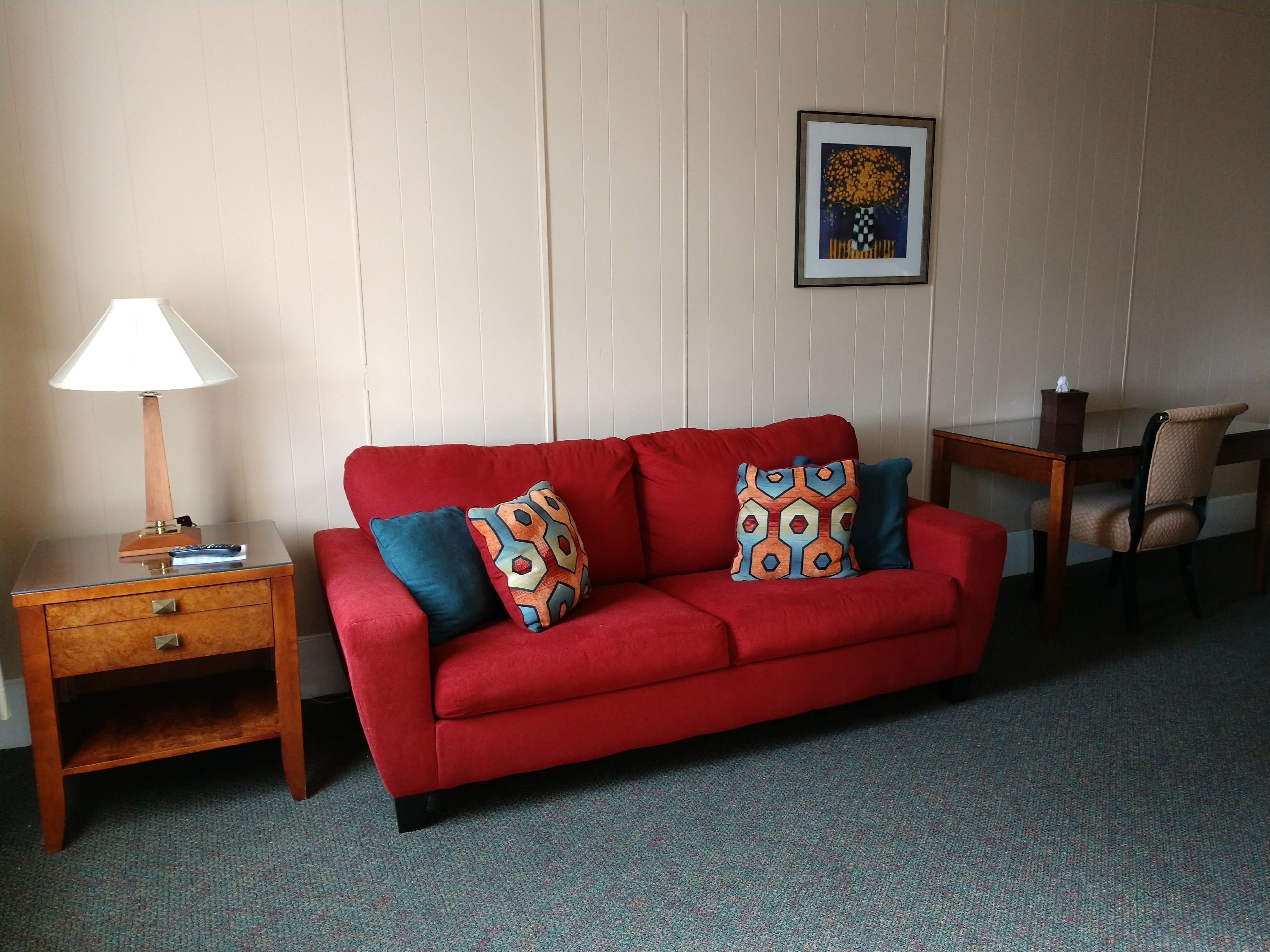A red sofa with patterned cushions, a wooden nightstand with a lamp, and a framed picture on the wall.