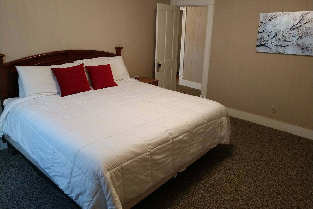A neatly made bed with white and red bedding in a room with beige walls and a carpeted floor.