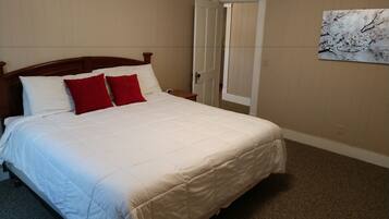 Una cama bien tendida con ropa de cama blanca y roja en una habitación con paredes color beige y piso alfombrado.