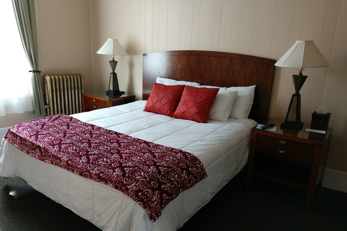 A hotel room with a large bed, two bedside tables, two lamps, a radiator, and a window with curtains.