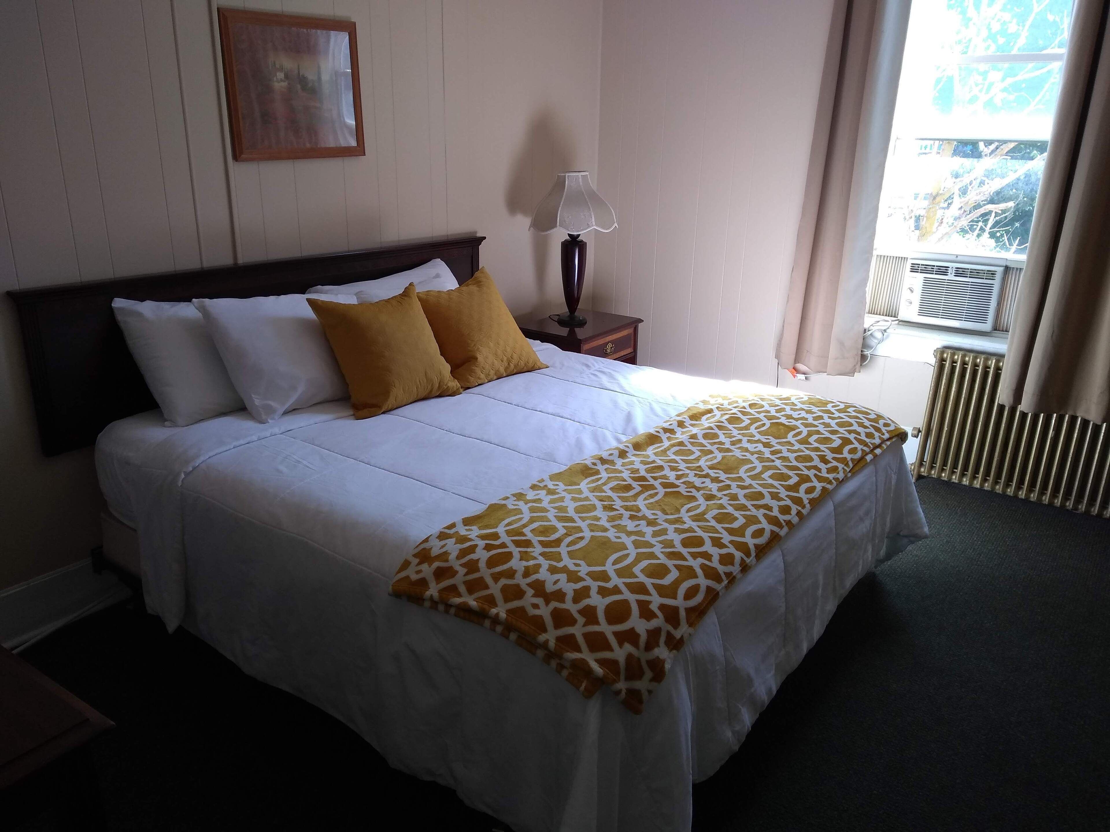 Una cama bien hecha con ropa de cama blanca y amarilla, una mesita de noche con una lámpara, y una ventana con cortinas.