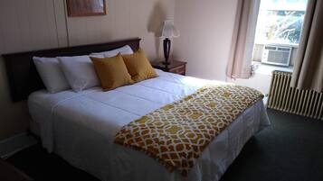 Una cama bien hecha con ropa de cama blanca y amarilla, una mesita de noche con una lámpara, y una ventana con cortinas.